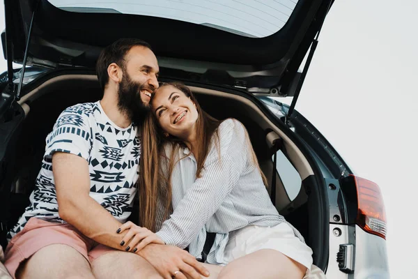 Happy middle-aged couple making stop while on a road trip, caucasian cheerful woman and man sitting in trunk of SUV car and smiling