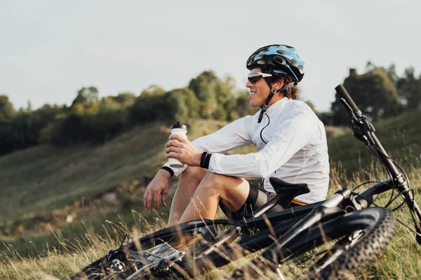 Professional Male Cyclist Drinking Water from Bottle, Man Sitting Near Bicycle During His Journey Outdoors in Countryside