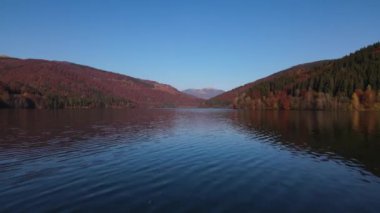 Drone, güneşli bir sonbahar gününde su yüzeyinde hızla ilerler, ufuktaki yüksek dağ ile dağların arasındaki gölü manzaralı bir şekilde görür.