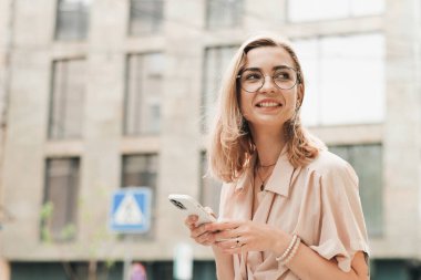 Gözlüklü Genç Kadın Ellerinde Akıllı Telefonu Tutuyor ve Gülümsüyor, Zarif Sarışın Kız Açık havada Uzaklara Bakıyor