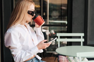 Akıllı Telefon Kullanırken Bir Kafe Masasında Oturan ve İçen Zarif Kadın, Güneş Gözlüğü Giyen ve Beyaz Gömlekli Kadın Kahve Dükkanı Terasında Buluşmayı Bekliyor