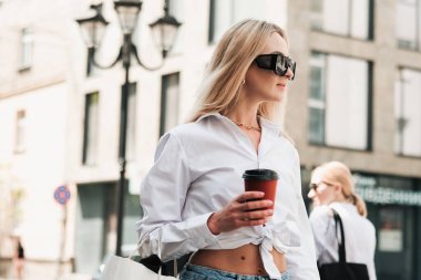 Şık Sarışın Kadın, elinde kahve fincanı olan, kendinden emin, güneş gözlüğü takan ve Altın Mücevher takan, Şehir Caddesinde yürüyen