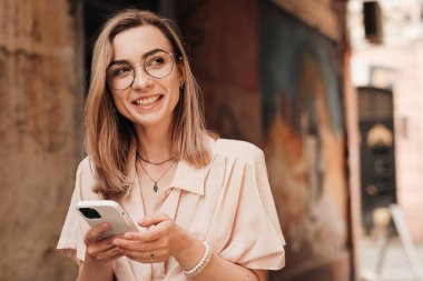 Gözlüklü Mutlu Genç Kadın Şehirde Yürürken Akıllı Telefonla Mesajlaşıyor, Gözlüklü Sarışın Kız Cep Telefonu Kullanıyor ve Gülümsüyor