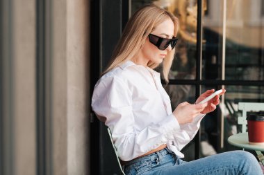 Kafe Masası 'nda oturan Kafkasyalı Kadın, Akıllı Telefon' da Mesajlaşan, Güneş Gözlüğü ve Beyaz Gömlek Giyen, Buluşmayı Beklerken Kendinden emin Kadın Girişimci