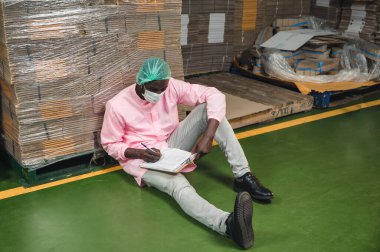 Steril üniformalı Afrikalı erkek envanter kontrol müdürü içecek işleme tesisinde depolanmış karton kutu siparişini kontrol ediyor.