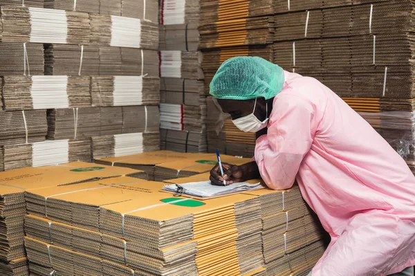 African man inventory control manager in sterile clothing checking order of cardboard box package stacked in warehouse at beverage processing plant