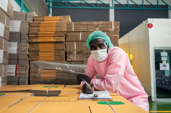 African man inventory control manager in sterile clothing checking order of cardboard box package stacked in warehouse at beverage processing plant