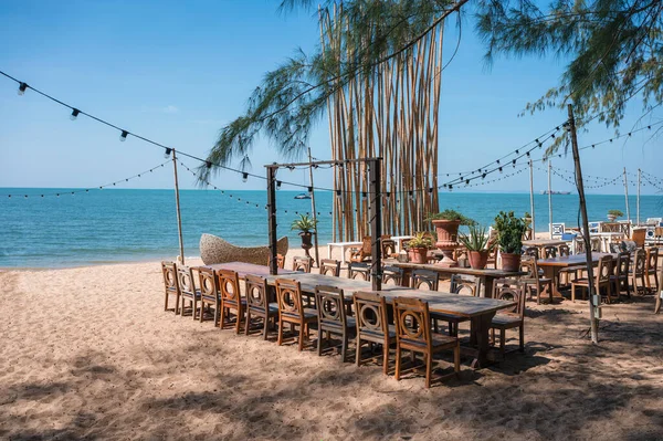 Celebration venue with wooden long table, chair, light bulb hanging and plant decoration in retro style on the beach in tropical sea