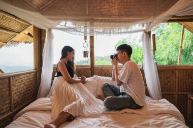 Genç Asyalı çift, tatilde doğanın arasında, ahşap barakadaki yatağın üzerinde eski bir kamerayla fotoğraf çekiyorlar.