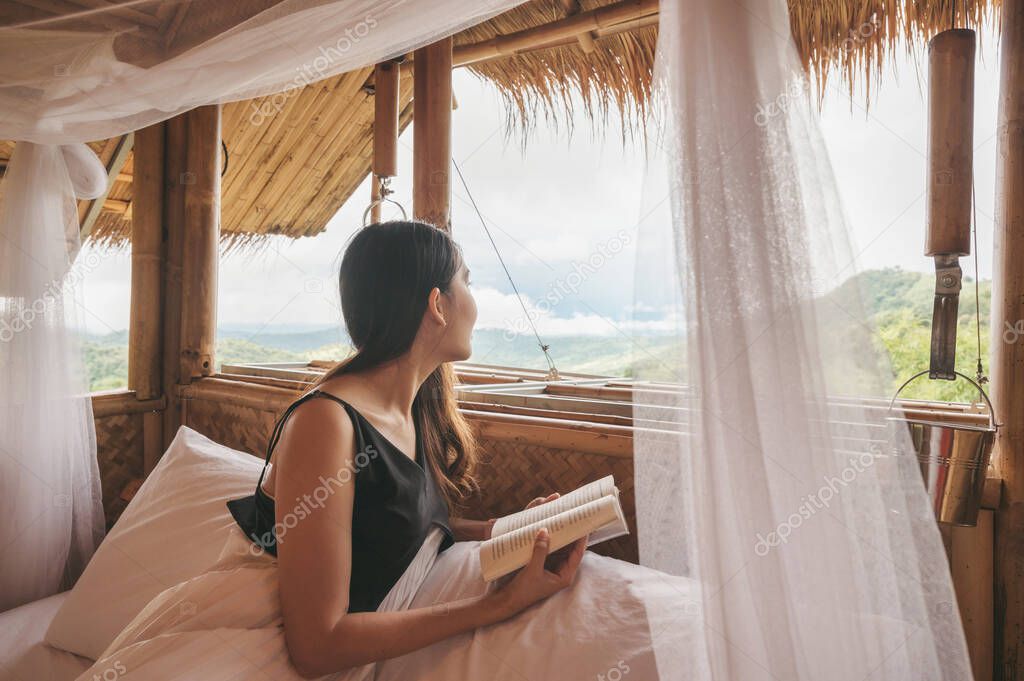 Beautiful young asian woman resting and reading a book on the bed in thatched hut among the mountain on vacation