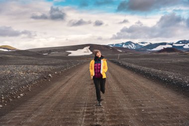Sarı ceketli Asyalı kadın İzlanda 'nın dağlık bölgelerinde Landmannalaugar' da toprak yolda koşuyor. 