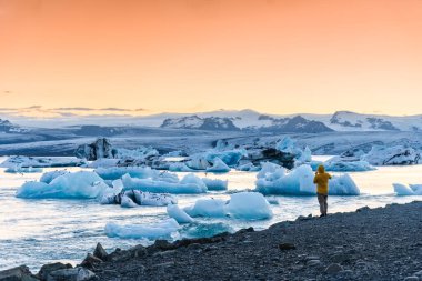 Ceketli turist Jokulsarlon Buzul Gölü, Vatnajokull Ulusal Parkı, İzlanda 'da gün batımında buzdağının keyfini çıkarıyor.