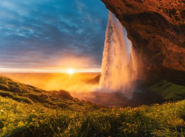 Güzel Seljalandsfoss şelalesinin manzarası ve yaz aylarında İzlanda 'nın güneyinde çiçek bahçesinde parlayan gece yarısı günbatımı.