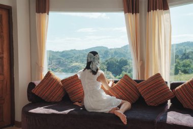 Hoş Asyalı kadın yaz tatilinde panorama odasındaki kanepede rahatlıyor ve tropikal orman manzarasının tadını çıkarıyor.