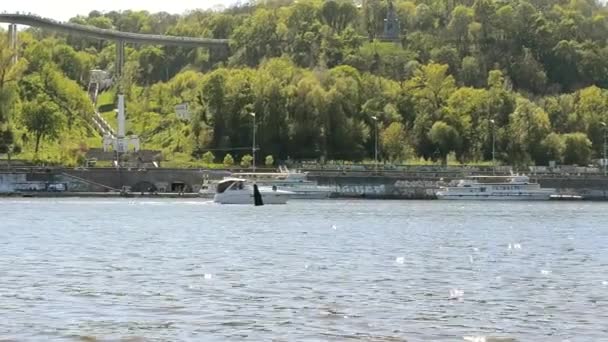 O barco navega ao longo de um grande rio em um dia ensolarado de primavera, vista lateral. O barco a motor se move rapidamente ao longo do rio, vista da praia. Edifícios da cidade ao fundo — Vídeo de Stock