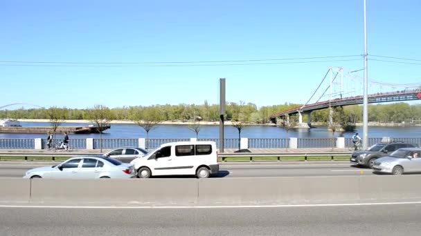 Киев, Украина - 24 апреля 2021 года: Автомобили едут по набережной автомагистрали. На заднем плане по реке плавает грузовая баржа. Движение по оживленной дороге в городе. — стоковое видео