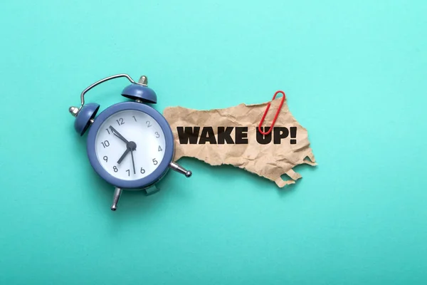 Text wake up on a piece of paper near an alarm clock on a blue background — Stockfoto