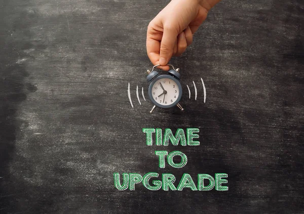 Man holds ringing alarm clock over words Time to Upgrade against chalkboard closeup. Changing to reach goals. Business development and innovation