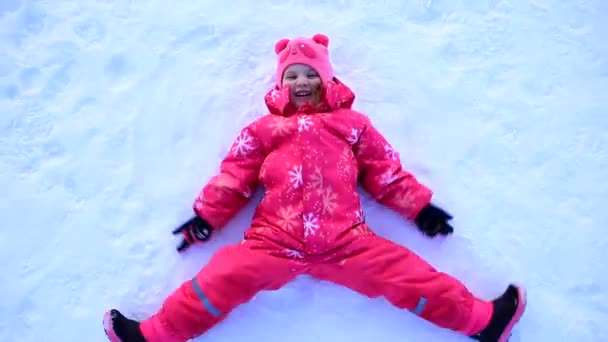 En ung flicka i gul jacka ligger i snön och gör en snöängel. — Stockvideo