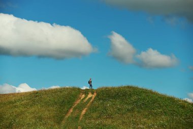 Yeşil bir tepenin üstünde duran kız turist ve mavi gökyüzünde kabarık bulutlar.