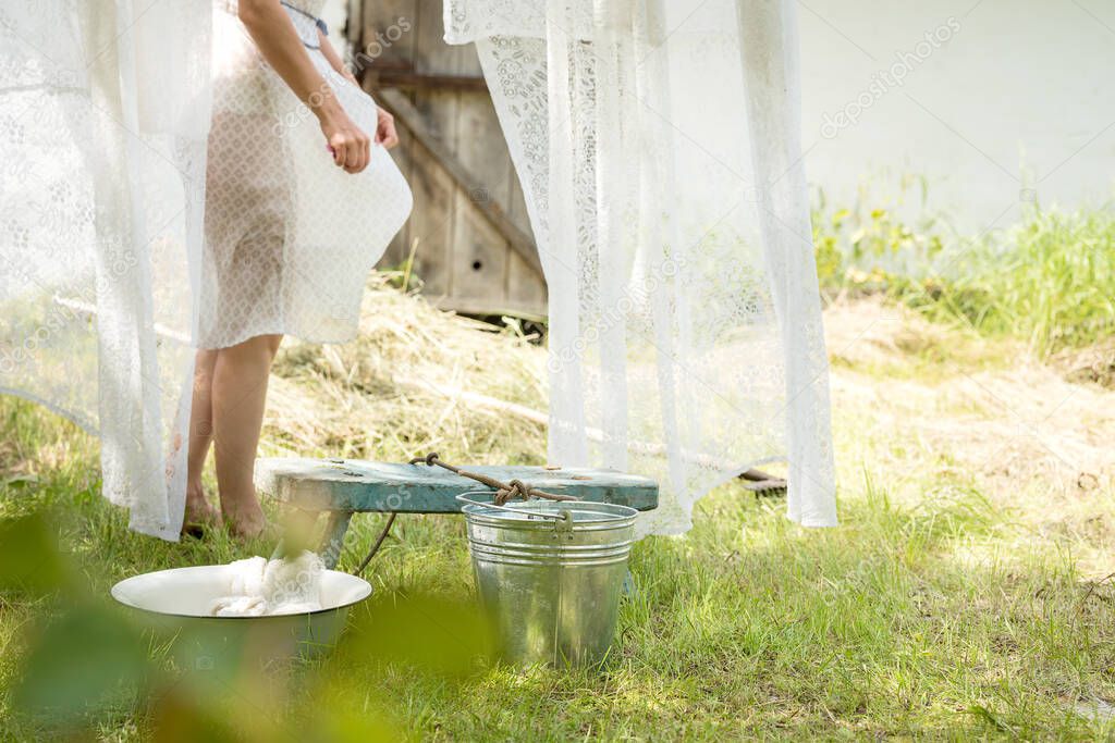 An artistic photo in a retro rustic style. A girl in a simple dress hangs the washed curtains in the garden. Laundry in the village.