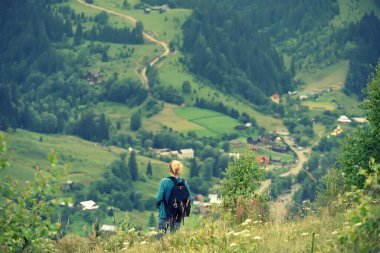 Sırt çantalı bir kız yeşil dağlara ve kırlara bakan bir açıklığın kenarında duruyor. Yaz dağlarındaki bir yürüyüşün atmosferi. yeşil turizm