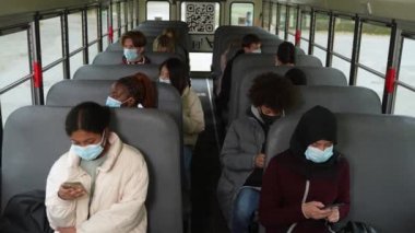 Okul otobüsünde farklı maskeli öğrenciler telefon kullanıyor.