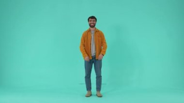 Kollarını göğsünde kavuştururken gülümseyen, gözlüklü, rahat giyinen bir erkek. Mavi arka planda tek başına dururken doğru tercihe güvendiğini ifade eden pozitif bir adam.