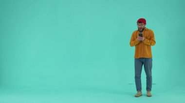 Örgü şapkalı ve gözlüklü neşeli bir erkek akıllı telefona ve mavi arka plana bakıyor, online mağazaların teklifinden etkilenmiş. Mutlu adam cep telefonundan tek tıklamayla alışveriş yapıyor.