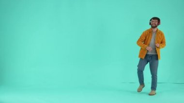 Gözlüklü ve kulaklıklı neşeli bir erkek mavi stüdyo arka planında izole edilmiş müzik ritmine ayak uyduruyor. Olumlu sakallı adam müziği seviyor ve dans ediyor.