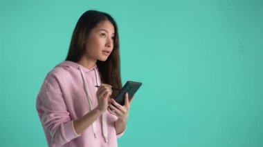 Stylus 'la cep telefonunda çalışan düşünceli genç bir kadın. Mavi stüdyo arka planında izole edilmiş bir fikri düşünürken kaşlarını çatıyor ve gülümsüyor. Akıllı telefondaki iş sırasında oldukça düşünceli bir kadın.