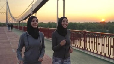 Sırıtan tesettürlü kadınlar ve spor giyimli koşucular şafakta nehir boyunca yaya köprüsünde koşuyorlar. Sportif Arap koşucular sabah sporunun keyfini çıkarıyorlar, aktif sağlıklı şehir sporları yaşam tarzı konsepti