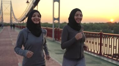 Şafak vakti yaya köprüsünde koşarken konuşan iki spor müslim bayanın yakın çekimi. Ortak sabah idmanı sırasında sağlıklı zinde Arap koşucuları
