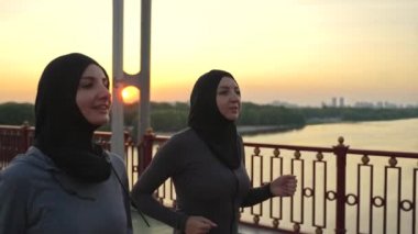 Gün batımında yaya köprüsünde koşarken spor kıyafetleri giyip konuşan pozitif Arap kadınlarının yakın çekimi. Uygun Müslüman koşucular ortak spor aktivitesi sırasında konuşmanın keyfini çıkarıyorlar.