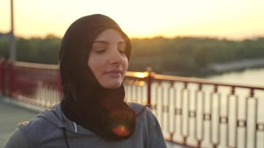 Şafak vakti sabah kardiyo antrenmanı sırasında tesettürlü güzel formda bir bayan koşucunun yakın plan portresi. Sportif Müslüman kadın yükselen güneş ışınlarıyla şehir köprüsünde koşuyor