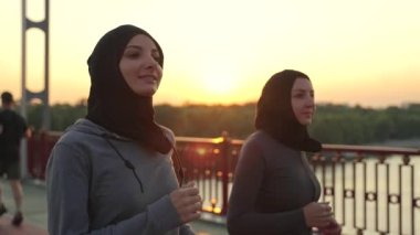 Gün batımında yaya köprüsünde koşan spor elbiseli Müslüman kadınların yakın çekimi. Sağlıklı Arap koşucular akşamları ortak eğlence aktivitesi, spor yaşam tarzı konsepti