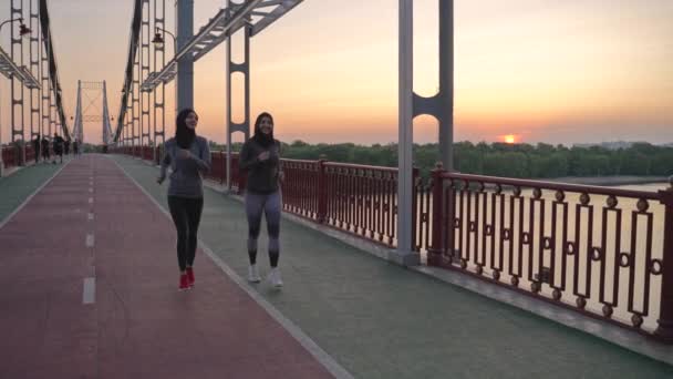 ヒジャーブとスポーツウェアを着た2人の健康なアラブ女性が街の歩道橋で夜のジョギングを楽しんでいます アクティブなイスラム教徒のランナーは 屋外で心臓トレーニングを行う余暇を過ごす — ストック動画