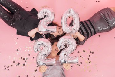 Bir grup kız arkadaş bir Noel partisinde eğleniyor. Pembe bir arka planda 2022 numaralı balonlarla yatıyorlar..