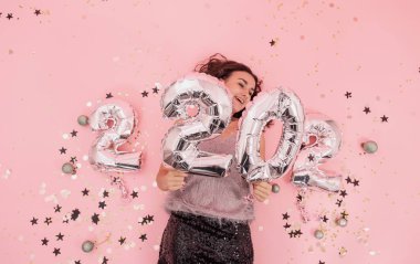 Pembe arka planda kıvırcık saçlı, neşeli, esmer, güzel bir kız. 2022 numaradan gümüş balonlar. Yeni yıl partisi konsepti..
