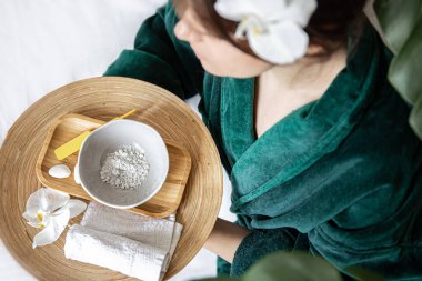 Yeşil bornozlu bir kadın elinde kil maske ve orkide çiçeğiyle kaplıcaya gider..