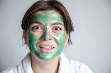 Yüzünde yeşil kozmetik bir maske ve gri arka planda beyaz bir bornoz olan çekici genç bir kadın. Evdeki spa tedavisi kavramı, uzay kopyası..