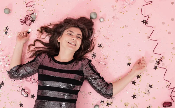 欢快的黑发女孩躺在粉红的背景上 与五彩纸屑和流苏在一起 这是庆祝圣诞节和新年的概念 — 图库照片