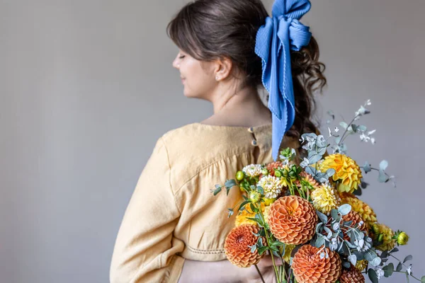 黄色とオレンジの菊の花束 灰色の背景を保持し 彼女の髪に青いリボンと黄色のドレスの若い女性 — ストック写真