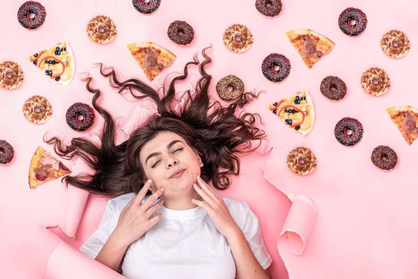 Приваблива молода жінка на рожевому тлі з піцою та пончиками, вид зверху . — стокове фото