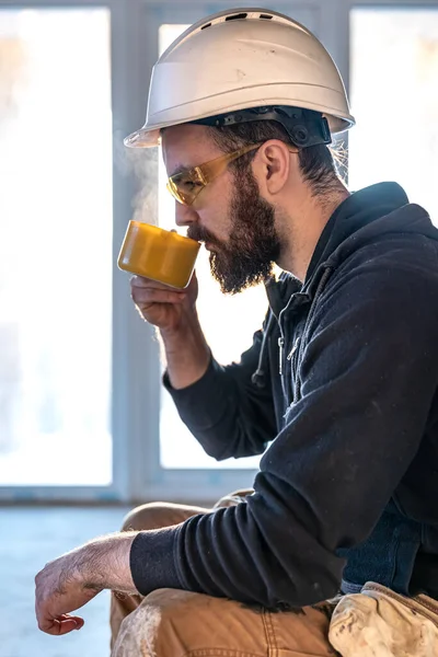 Un hombre constructor en un casco y gafas bebe una bebida caliente. — Foto de Stock