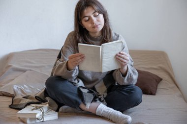 Genç kadın yatakta otururken bir kitap okuyor..
