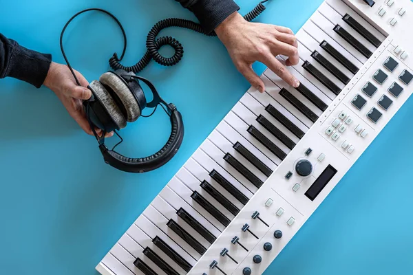 Чоловічі руки грають музичні клавіші і тримають навушники на синьому фоні, вид зверху . — стокове фото