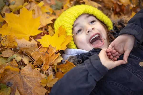Egy sárga kalapos kislány fekszik az őszi lombozatban.. — Stock Fotó