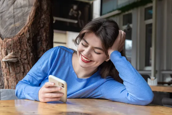 Una joven con auriculares inalámbricos usa el teléfono mientras está sentada en un café. — Foto de Stock