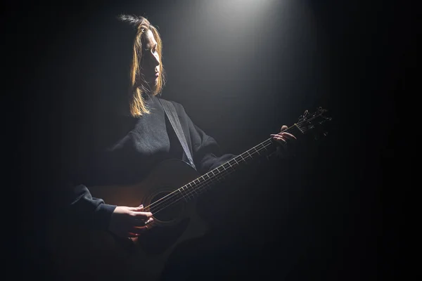 Meisje met een akoestische gitaar in het donker onder een lichtstraal. — Stockfoto